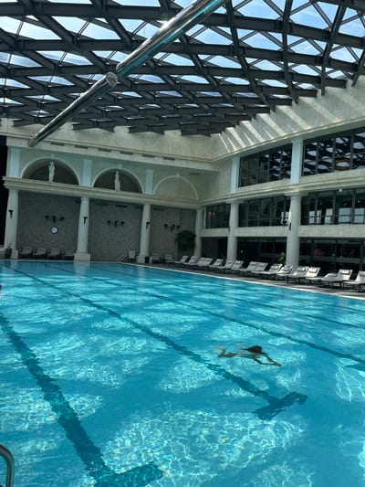 The image shows an indoor swimming pool with a woman swimming. The pool is large and rectangular, with lane lines visible on the bottom. The water is clear and blue, with sunlight reflecting off the surface. To the left of the pool is a building with a large glass and steel structure, and a row of white lounge chairs. The architecture suggests a modern hotel or resort. The lighting indicates it is daytime. The woman is swimming freestyle and appears to be alone in the pool.