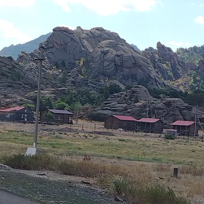 The image depicts a rural landscape in the vicinity of Toraygyrov, Kazakhstan, characterized by rocky hills and a small cluster of buildings. The terrain is varied, with rugged, grey rock formations dominating the background, suggesting a mountainous or hilly region. The foreground features a dry, grassy field with scattered vegetation, indicative of a semiarid or steppe environment.
Several small, wooden houses with red roofs are visible, situated in a line and separated by fencing. These structures appear to be residential or perhaps part of a small settlement. The houses have distinct window and door features, and one structure in the center appears to be a shed.
A prominent feature in the mid-ground is a utility pole with wires, suggesting the presence of infrastructure in the area. The overall scene conveys a sense of remoteness and a simple, rural lifestyle. The sky is a pale blue with scattered white clouds, suggesting fair weather.