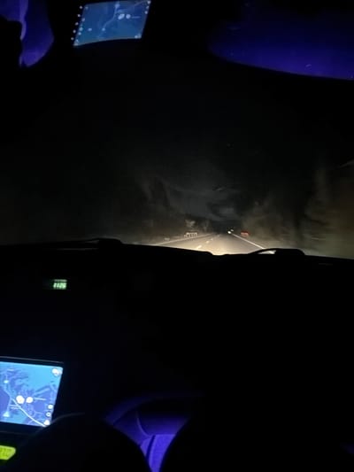 The image depicts the interior of a vehicle at night. Through the windshield, a road stretches ahead, illuminated by the vehicle's headlights. There is another vehicle with a red taillight visible on the road ahead. Inside the car, two dashboard screens are visible. One screen displays a map. The interior is dark with hints of light reflecting off surfaces. It cannot be definitively determined if this is Mezdreya, Bulgaria from the image.