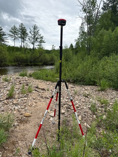 An outdoor, eye-level shot captures a surveying instrument, likely a GPS receiver, mounted on a tripod in the foreground. The instrument is positioned on a rocky, gravelly riverbank with scattered green vegetation. Behind the instrument, a body of water, a river, flows gently. The background is dominated by lush green trees and foliage, with a cloudy, overcast sky above. The location is identified as Murtygit, Russia. The scene is static and shows no people or any ongoing activity.