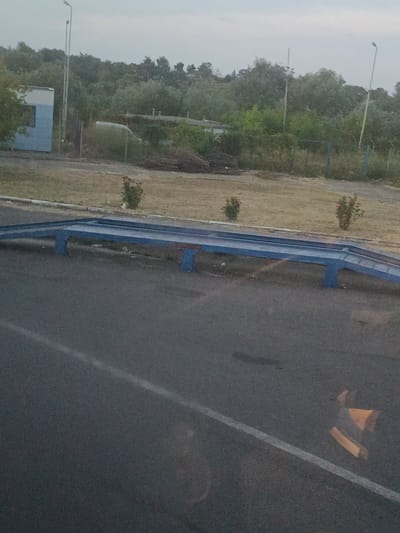 The image depicts an outdoor scene in Oancea, Romania, focusing on a blue metal structure with ramps, likely a weighing station or a platform for loading/unloading. The structure is situated next to a paved road with a white line marking. Behind the structure, there is dry grass and some sparse vegetation, including two small rose bushes. In the background, a dense line of trees forms a natural barrier. Further back, a light blue and white building with a dark rectangular opening is visible, alongside utility poles and what appears to be a fence. The sky is overcast. No individuals are present in the frame.