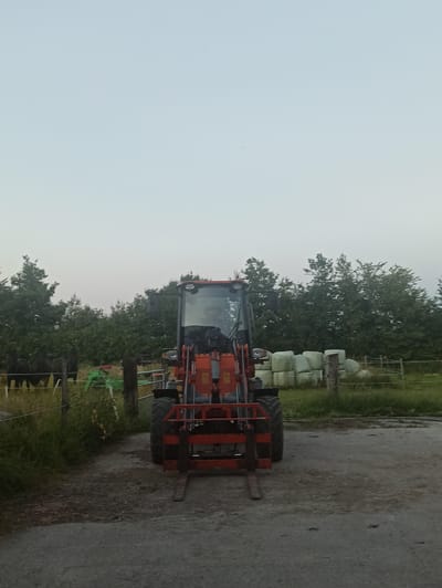An orange front loader tractor is parked on a gravel surface in front of a field containing stacked hay bales wrapped in white plastic. In the background, a line of trees separates the field from the sky. A fence runs along the edge of the field. A small green tractor is visible in the distance to the left of the front loader. The sky is overcast and light. There are no people visible in the image. The scene appears to be in a rural setting.