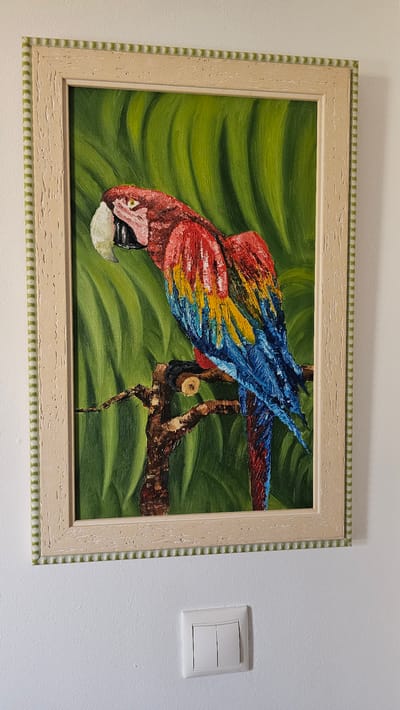 The media file depicts a framed oil painting of a scarlet macaw perched on a branch. The background is a vibrant green with a wavy pattern, suggesting foliage. The macaw is painted with thick, textured brushstrokes, showcasing its bright red, yellow, and blue feathers. Its head is tilted downwards, and it appears to be looking towards the viewer. The frame of the painting is light-colored wood with a decorative border. To the right of the painting, on a white wall, is a white light switch. The scene appears to be indoors, likely in a home or gallery setting. There is no visible text in the image. The setting suggests a still-life arrangement, with the focus on the artwork and its immediate surroundings. The bright colors of the parrot and the background evoke a sense of tropical vibrancy.