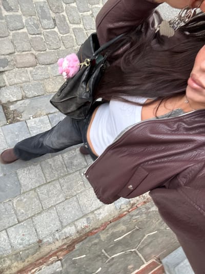 A high-angle, downward-looking photograph captures an individual on a paved street in Tielrode, Belgium. The person, identifiable by long, dark brown hair, wears a white ribbed tank top, dark grey denim jeans, and brown flat shoes. A dark reddish-brown jacket with a visible zipper is worn. A black leather shoulder bag is slung over the left shoulder, adorned with a bright pink, fluffy keychain. A silver necklace with a small pendant is visible on the chest. Near the right shoulder, a hand, largely out of frame, holds a set of keys with multiple silver keyrings. The ground surface consists of irregular grey cobblestones, with a lower edge of reddish-brown bricks and grey concrete/mortar visible along the right side of the frame.
