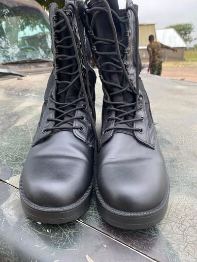 Two black leather military boots are centered on a dirty, mottled metal surface. The boots are laced and feature black nylon fabric sections along the shaft. A blurred figure in camouflage attire stands in the background near a building with a white roof. A car windshield and foliage are visible reflecting on the metal surface beneath the boots, suggesting they are resting on a vehicle. The sky is overcast.