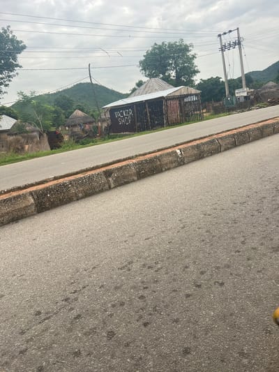 The image shows a rural scene in Kona Garu, Nigeria. A paved road runs diagonally through the frame, with a concrete and stone curb separating it from a grassy area. On the left side of the road, there are several buildings. One building has a corrugated metal roof and appears to be a shop, with the text "TOKIZA SHOP" painted on its side. Next to it are traditional thatched-roof huts. Beyond the buildings, a lush green hillside rises, dotted with trees. Overhead, power lines stretch across a cloudy sky. The overall impression is of a quiet, everyday scene in a developing community. The lighting suggests it is daytime, possibly overcast.