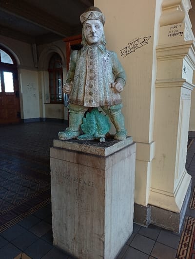 The image displays a weathered statue, identified as "GRÜN WENZEL," situated indoors on a rectangular stone pedestal. The statue depicts a male figure, appearing as a boy or young man, wearing a hat, a long-sleeved, buttoned coat, breeches, and boots. His face exhibits discoloration, with a prominent white substance around the nose and mouth area. The figure stands with legs spread, hands clenched into fists at his sides. Positioned between his legs, at groin level, is a green, leaf-like or plant-like object, designed as a spouting element. The pedestal supporting the statue is carved with the inscription "GRÜN WENZEL" on its visible front face.
The statue is located within an indoor public space, possibly a hall or covered arcade. To the left, a partially open arched wooden door allows ambient light, revealing an exterior or adjacent brightly lit area. Beyond this, another arched opening leads deeper into the structure. To the right of the statue, a cream-colored architectural pilaster features black marker graffiti, including the legible text "LEANOX IST EIN PENNER!!". The floor is covered with patterned tiles, exhibiting geometric designs in dark grey, brown, and possibly reddish hues. A dark, paneled ceiling is visible overhead. This scene is located in the city of Altenburg, Germany.