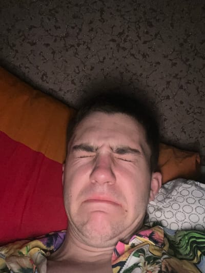 The image shows a close-up of a person's face, with their eyes squeezed shut and their mouth contorted in what appears to be a grimace or expression of discomfort. The person is lying down, with pillows and a dark, textured surface visible in the background. The setting appears to be indoors, possibly a bedroom. The person is wearing a brightly colored, patterned shirt. The image does not provide any information about the city of Ponebel, Ukraine, or any specific location within it.