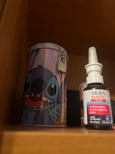 The image shows a close-up of objects stored on a shelf. In the foreground, there is a metal tin with a cartoon character, Stitch, printed on it. The tin has a padlock attached to its lid, suggesting it might be a savings tin or a keepsake box. Behind the tin, a bottle of nasal spray is visible, with text in Cyrillic script. The label reads "AKBA MAPIC" and mentions it is a spray for nasal congestion. The background consists of a plain brown surface, likely the interior of a cabinet or shelf. The lighting is dim, indicating an indoor setting, possibly a room in a home or office. There are no people or any specific time-of-day cues. The overall impression is that of personal items stored away.