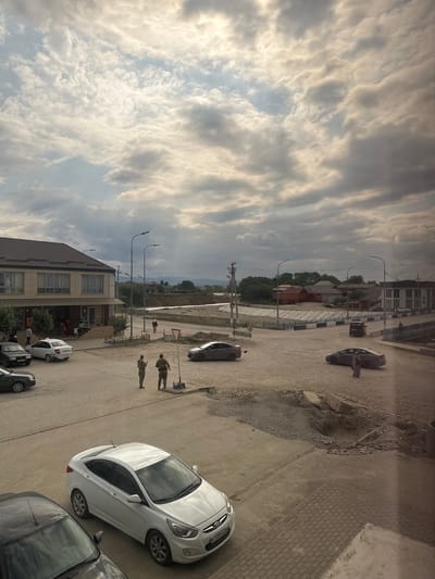 The image depicts a view of Achkhoy-Martan, Russia, on a cloudy day. In the foreground, a white sedan is parked, and partially visible is the front of a dark-colored car. A few other cars are parked and driving on the street. Two individuals dressed in military-style uniforms are standing on the side of the road, near a red and white triangular yield sign. To the left, a two-story building with a brown roof houses a shop with several windows. People are seen entering and exiting the shop. In the middle ground, a river or canal runs through the scene, with a bridge crossing it further down. Buildings with red and white roofs are visible on the opposite bank, along with trees and distant hills. The road surface appears to be a mix of asphalt and gravel, with some areas of construction or disrepair. The sky is filled with dramatic, heavy clouds, with beams of sunlight breaking through in places.