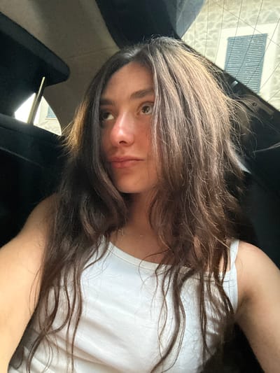 The image features a young individual, likely female, with long, dark brown hair, seated inside a vehicle. The subject is wearing a white sleeveless top and a delicate, gold-colored chain necklace with a small circular pendant. Her gaze is directed upwards and towards the left side of the frame. The interior of the vehicle is dark, with elements such as a headrest and roof lining visible. Through the rear window, a light-colored building exterior is discernible, featuring a window with distinct horizontal slatted blinds. The scene is consistent with a daytime setting in the city of Deià, Spain.