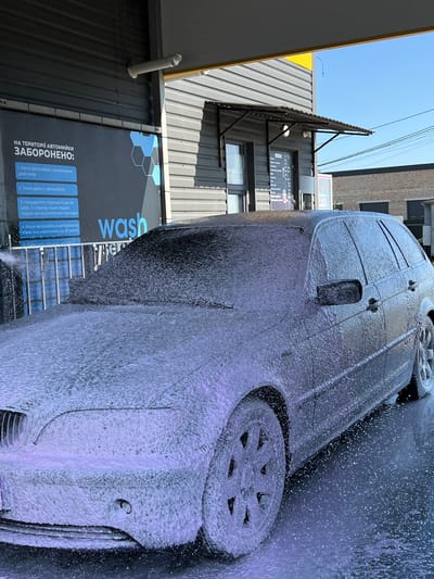 A dark-colored car, covered in white foam, is parked at a self-service car wash. The car appears to be a station wagon, with its windows, hood, and roof obscured by the suds. Water droplets are visible on the ground, suggesting the washing process is underway. The car wash structure features a corrugated metal exterior and a yellow awning above the entrance. A blue and white sign on the wall behind the car lists rules in Ukrainian, including prohibitions against washing a car with the engine running, washing the engine, and directing water at people or electrical equipment. The scene is set outdoors under a clear blue sky.