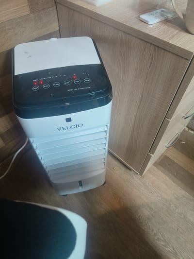 In the city of Redkino, Russia, a white and black evaporative cooler with the brand name VELGIO is situated on a wooden floor next to a light brown wooden cabinet. The cooler has a control panel with buttons labeled "POWER," "ON/OFF," "SPEED," "TIMER," "MODE," "COOL," and "SWING." Several red indicator lights are illuminated on the panel. A white power cord is visible extending from the cooler. On top of the cabinet, a white remote control with several buttons and a light-colored mug are present. The environment appears to be indoors, with the wooden floor and cabinet suggesting a domestic or office setting.