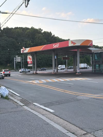 The image depicts a self-service gas station in Pocheon-si, South Korea. The gas station has an orange and white canopy with "Self" written on it in large white letters. An SK logo is prominently displayed on a pole under the canopy. Several cars are visible on the road and at the gas station, including a white SUV and a dark-colored SUV. In the background, there are trees and a building. The scene appears to be during daylight hours, with a clear sky and some clouds. The overall atmosphere is that of a typical urban or suburban environment.