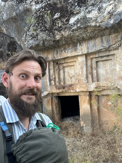 A man with a beard and wide eyes takes a selfie in front of ancient rock-cut tombs in the city of Seydikemer, Turkey. The tombs are carved into a rocky hillside, with a dark opening visible in the center. The man is wearing a striped shirt and a vest, and has a backpack over his shoulder. The environment appears dry and arid, with sparse vegetation. The image is taken in daylight.