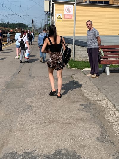 In a sunlit outdoor scene, likely a train station platform in Kungur, Russia, a group of people are gathered. The ground is paved with asphalt and gravel. In the foreground, a woman with dark hair, wearing a black crop top and a leopard-print skirt, walks away from the camera, carrying a black backpack. To the right, a man with short gray hair, wearing sunglasses, a striped t-shirt, and dark trousers, stands looking towards the left, holding a carton of juice. Behind them, several other individuals are visible walking along the platform, some carrying bags. A yellow building with a corrugated metal lower section runs along the right side of the platform, and a wooden bench with metal legs is situated in front of it. Overhead, a complex network of electrical wires crisscrosses the sky. A yellow warning sign with a black border depicting a person and a triangle is mounted on a pole near the building.