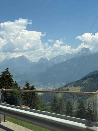 The image shows a view from a cable car, looking out over a mountainous landscape. The sky is a bright blue with fluffy white clouds. In the distance, large, jagged mountains rise up, some with snow-capped peaks. The lower slopes of the mountains are covered in dense green forests. Below the mountains, a town or village can be seen nestled in the valley, with buildings and roads visible. The foreground of the image is dominated by the metal railing of the cable car, which is silver and appears weathered. There are also some trees visible in the foreground, which are dark green and appear to be pine trees. The overall scene suggests a sunny day in a mountainous region, likely in Switzerland given the location context. The image conveys a sense of height and expansive natural beauty.