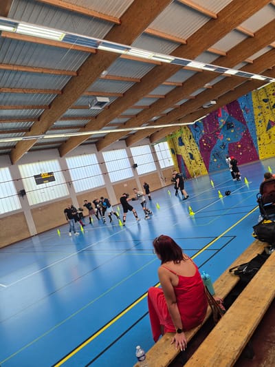 The image shows the interior of a sports facility, likely a gymnasium or multi-purpose hall, in Leers, France. A group of people are inline skating on a blue court marked with yellow lines and yellow cones. Some are lined up practicing, while others appear to be observing. In the background, a colorful climbing wall is visible. A woman in a red dress sits on a wooden bench in the foreground. The building's architecture includes a high ceiling with exposed wooden beams and rows of windows.