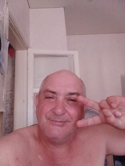 A close-up, selfie-style photograph shows a bald, middle-aged Caucasian man with fair skin and stubble. He is shirtless and looking directly at the camera, with his right hand making a peace sign near his eye. He appears to be indoors, with a white door frame and a partially visible, frosted glass pane in the background. To the left, there is a glimpse of what appears to be a wall with some objects hanging on it, possibly clothing or a tapestry. The lighting is soft and diffuse. There is no indication of the location being specifically Krasny Yar, Russia, or any other city.