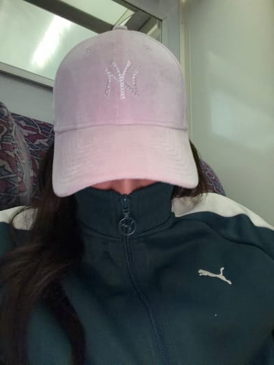 A person wearing a light pink baseball cap with a sparkly "NY" logo is visible. The cap is tilted towards the camera. The person is also wearing a dark green Puma jacket with white trim. Their dark hair is seen below the cap. The setting appears to be indoors, possibly a vehicle due to the visible window frame. A patterned seat or cushion is also visible.