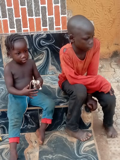 Two young children are seated side-by-side on a patterned tiled surface, possibly outdoors on a porch or veranda. The child on the left is younger, appearing to be a toddler or very young child, with dark skin and hair styled in small braids close to the scalp. This child is bare-chested and wears light blue pants that are rolled up at the cuffs, revealing a red lining. They are holding a tangled white cord or a pair of white earphones in their hands, looking slightly to their right with a neutral expression. Small hoop earrings are visible in their ears.
To the right sits an older child, also with dark skin, who has a completely shaved head. This child is wearing a long-sleeved orange shirt that appears somewhat worn, with visible tears or holes around the shoulder and upper arm area. They are also wearing dark-colored pants. This child is looking off to their right, away from the camera, with a pensive or neutral expression.
Both children are barefoot. They are sitting on a bench-like structure or a raised platform covered with tiles featuring a swirling, marbled design in shades of dark blue, black, white, and beige/brown. The background consists of a wall. The lower part of the wall continues the marbled tile pattern, while the upper part features a brick-like pattern in alternating rectangular shapes of orange, gray, and white. To the far right, another wall with a lighter, textured, somewhat embossed pattern is visible. The lighting suggests it is daytime. No text is visible in the image.