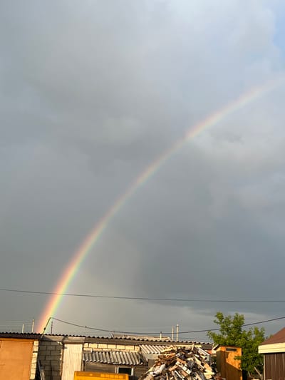 A faint rainbow arcs across a cloudy, overcast sky above a collection of low-rise buildings and a pile of chopped wood in what appears to be a rural or suburban area. The buildings have corrugated metal roofs and some are constructed from concrete blocks. A small wooden outhouse is visible near a leafy green tree. Electrical wires are strung across the scene, indicating a settlement. The lighting suggests it is late afternoon or early evening. The location is stated to be in Timiryazev village district, Kazakhstan. There are no people visible in the image.