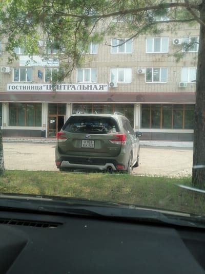 The image captures a rear view of a green Subaru Forester parked in front of a brick building with a sign reading "ГОСТИНИЦА ЦЕНТРАЛЬНАЯ" (Hotel Centralnaya). The building appears to be a multi-story structure with numerous windows and air conditioning units. A tree with green foliage partially obscures the view of the building. The car has a license plate that reads "O 938 XK 125 RUS". There are stickers on the rear window of the car, one with text that appears to be in Russian and includes a phone number. The foreground shows a portion of a car's dashboard and windshield, suggesting the photo was taken from inside another vehicle. The ground is paved and there is a strip of grass in front of the building. The setting appears to be a commercial area in the city of Lesozavodsk, Russia.