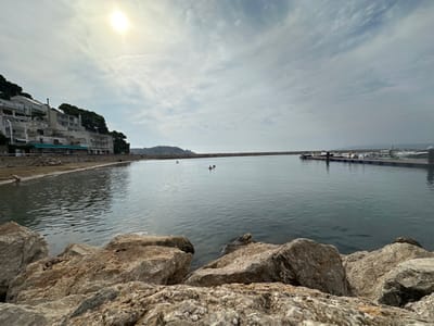 The image captures a coastal scene in L'estartit, Spain, from a low perspective, overlooking a sheltered bay or harbor. The foreground is dominated by large, irregular, light-colored boulders. The central area features a calm body of grey-blue water. Two individuals are observed in the water, positioned towards the left-center, appearing to swim or float. Along the left shoreline, a narrow sandy strip abuts a retaining wall, behind which a series of multi-story, light-colored buildings with green awnings are situated on a tree-covered incline. To the right, a lengthy, low rock breakwater extends into the water, sheltering a marina. Several white boats are moored at floating docks within this area. One individual is discernible standing on the dark pier section of the marina. The sky is partially cloudy, with the sun visible as a bright disc in the upper-left, causing a glare on the water's surface. Distant landmasses are visible on the horizon.