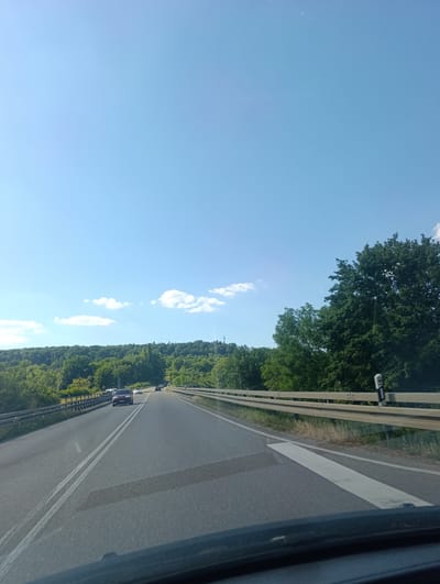 This media file depicts a view from a vehicle driving on a highway in Neckartailfingen, Germany. The scene is a bright, sunny day with a clear blue sky dotted with a few white clouds. The highway, which appears to be a two-lane road with double white lines marking the lanes, is bordered by a metal guardrail.

To the left and right of the highway, lush green trees and dense foliage form a picturesque backdrop, indicating a rural or semi-rural setting. In the distance, a rolling hill covered in trees is visible. A few cars are driving on the highway, suggesting moderate traffic.

The time of day is clearly daytime, likely mid-morning or early afternoon, given the bright sunlight and the shadows cast. The weather appears to be pleasant and warm. There are no discernible people visible as subjects, but the presence of vehicles indicates their activity in traveling.

Notable details include the white markings on the road, which include a large arrow painted on the right side of the lane, likely indicating a lane change or merging point. A white post with some markings is also visible on the right side of the road, possibly a road marker or reflector. The overall impression is one of a scenic drive through a green landscape on a beautiful day. No text is clearly visible in the image.