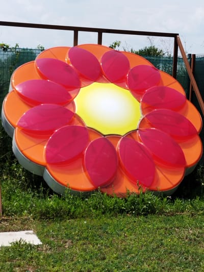 A large, colorful, abstract flower sculpture made of plastic is displayed outdoors on a grassy area in Clinceni, Romania. The sculpture features bright orange petal-like shapes layered with translucent pink petals. The center of the flower is a yellow disc with a gradient of light. The sculpture is supported by a dark metal frame structure in the background. The environment includes lush green grass, some scattered weeds, and a cloudy sky. A portion of a light-colored concrete slab is visible in the lower left corner of the image, partially obscured by grass.
