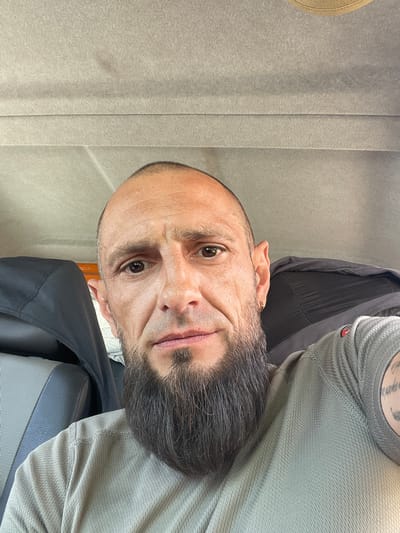The media file is a close-up, eye-level, portrait-style photo of a man's face taken from a slightly low angle. The man has a bald head with graying hair around the sides and a full, dark beard with some gray mixed in. He has brown eyes and is looking directly at the camera with a slight smile or smirk. He is wearing a light gray, long-sleeved shirt.
The background is out of focus but appears to be an indoor setting with textured gray walls or fabric. There is a hint of a black object behind him, possibly a piece of furniture or clothing. The lighting suggests it is daytime, though no specific weather conditions are visible.
The man has tattoos visible on his neck and shoulder, but they are mostly obscured and difficult to read. There is a small gold earring in his left ear. The overall mood of the image is casual and direct. The provided location context is Vynnyky, Ukraine. No visible text is present in the image.