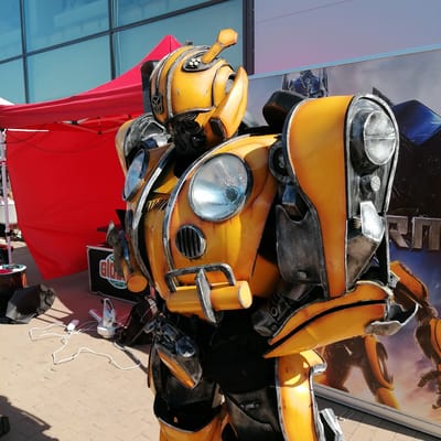 A detailed, full-body costume of Bumblebee from the Transformers franchise stands upright outdoors under bright sunlight in Yudino, Russia. The costume is primarily yellow with weathered dark gray and silver accents, mimicking battle damage and robotic parts. Its head features the iconic Autobot symbol on the left cheek, a dark eye visor, and an antenna-like protrusion on the top with small circular elements. The chest incorporates two large, ribbed circular headlights and a small rectangular grille-like vent below the left headlight. Visible black under-suit material shows between the segmented yellow and gray armor plates on the torso, arms, and legs.
In the background, a modern building with large, reflective blue-tinted glass windows is visible on the left. A bright red pop-up canopy or tent, supported by white poles, is partially seen behind the costume on the left side. To the right, a large promotional banner displays imagery from the Transformers franchise, featuring a blue and red robot (Optimus Prime) on the left part of the banner and a yellow-orange robot (likely Bumblebee) on the right, partially obscured by the costume's leg. Stylized text, partially visible, includes Cyrillic characters.
The ground is paved with light-colored rectangular concrete pavers. In the foreground and midground, various items are present: a black fabric bag, a small dark stand with minor objects, a black equipment case displaying a "Globus" logo, and a white power cord snaking across the pavers. The cord leads to a white electronic device, possibly a fan or fog machine, positioned near the base of the costume. Distinct shadows of the costume and other objects are cast on the paved ground, indicating direct sunlight from the upper right. No other individuals are visibly present beyond the person inside the costume.