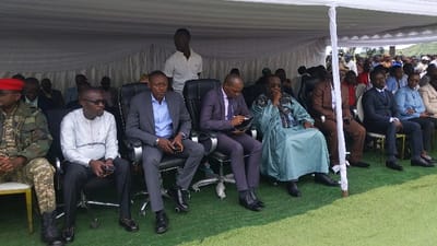 The image shows a group of men seated under a white tent on a field of artificial grass. The men are all dressed in formal attire, with some wearing suits and others wearing traditional clothing. They are seated in black chairs with a white support pole in the middle. One man is wearing a military uniform and a red beret. The location is the city of Bukavu, Democratic Republic of the Congo, at coordinates -1.624, 29.255. The men are seated attentively and appear to be participating in a formal event.