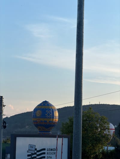 The image depicts an outdoor scene in Bartkowa-Posadowa, Poland, during what appears to be late afternoon or early evening. Dominating the foreground is a tall, metallic pole, slightly off-center to the right. Behind it, a large, inflatable hot air balloon, primarily blue with gold ornamentation, is prominently displayed. The balloon features intricate patterns, including what looks like a sun symbol and decorative banners. To the left of the balloon, a utility pole with various attachments is visible.

In the mid-ground, a sign with black and white stripes and text that reads "Droga Wewnętrzna" (meaning "Internal Road") is affixed to a building or structure. Below the balloon, on what appears to be a building facade, the text "LEMON RESORT SPA" is displayed. Further in the background, a lush, tree-covered hillside rises, with several cell towers visible on its crest. The sky is a pale blue with wispy white clouds, suggesting fair weather. The overall impression is that of a commercial establishment or resort area, likely a tourist attraction given the prominent hot air balloon.