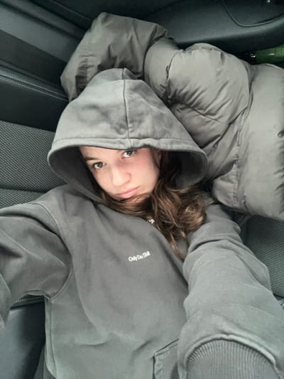 A young individual, appearing to be female, is depicted reclining within the interior of a vehicle, positioned at a slight angle with her head towards the upper left. She wears a dark grey hooded sweatshirt with the text "Only Do Still" printed in white on the chest; the hood is pulled over her head. Her medium-length brown hair is visible around her face and shoulders. Her gaze is directed towards the camera with a neutral expression. To her right, a grey, padded object, possibly a jacket or blanket, is visible. The vehicle's interior features dark grey upholstered seats with a discernible pattern and a visible seatbelt mechanism with light stitching on the upper left. The image lacks specific visual identifiers to determine a precise location within Stara Pazova Municipality, Serbia.