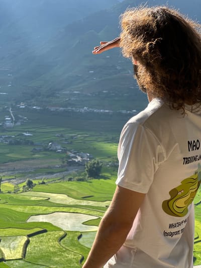 A person with long, curly, brown hair is viewed from behind, standing outdoors and looking into a vast valley in Tú Lệ, Vietnam. The individual, whose face is not visible, is wearing a white short-sleeved t-shirt. Their right hand is raised to shield their eyes from the sun. The t-shirt features text on the back: "MAO TREKKING AND" on the upper right side, and "WHATSAPP +84" and "Instagram: ma" (partially visible) towards the bottom right. An illustrated graphic depicting green terraced fields with small houses is also printed on the back.

The background reveals a expansive landscape characterized by lush green terraced rice paddies, some filled with reflective water, cascading down hillsides. In the midground, a small village with scattered houses and a winding road is visible along a river or stream. Distant mountains rise, creating a hazy, layered appearance under what appears to be bright sunlight from the left, highlighting the person's hair and shoulder.