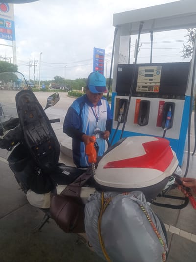 At a PTT Station in Tha Ruea, Thailand, a female employee in a blue uniform and cap fills a motorcycle with gasoline. The motorcycle, packed with a leather bag and a large grey bag secured with bungee cords, stands next to a gas pump with three fuel options. The station's price sign is visible in the background.