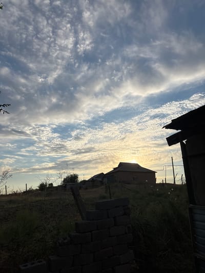 The image depicts a rural landscape at dawn or dusk, likely in the Жібек жолы ауылдық округі, Kazakhstan. The sky is filled with a dramatic display of scattered clouds, ranging from wispy cirrus formations to denser, more textured clouds, illuminated by the low-lying sun which is partially obscured by a building. In the foreground, to the right, a dark, silhouetted structure, possibly an outbuilding or shed, dominates the frame, with its roofline sharply defined against the sky. To the left and center, a rural dwelling with a dark roof is visible, set against the backdrop of rolling hills and sparse vegetation. A low, rough-looking wall constructed from grey bricks is in the immediate foreground, partially obscuring the view of the property. Barbed wire fences and thin, dry vegetation are scattered across the landscape, suggesting a dry, possibly arid environment. The overall impression is one of rustic simplicity and a serene, natural setting. There are no people or animals visible in the image.