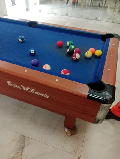A pool table is positioned on a light-colored tiled floor. The pool table has a blue felt surface with several pool balls scattered across it. The balls are of various colors and numbers, including red, blue, green, yellow, and purple. Some of the balls have visible numbers, such as "2", "6", "8", "9", "15", and "1". The pool table has a wooden frame with metal accents and black corner pockets. The background shows a reflective surface, possibly glass or a mirror, and blurred figures of people and furniture, suggesting an indoor setting like a bar or recreational area. The floor tiles exhibit a marble-like pattern. The scene appears to be located in the city of Kwale, Nigeria. There are no discernible actions or interactions between people and the objects.