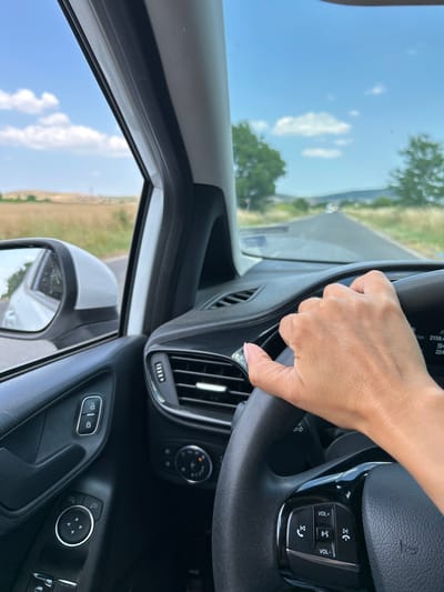 The image shows a close-up, interior view of a car's dashboard and steering wheel, with a person's hand holding the wheel. The hand, belonging to a woman with neatly manicured nails, is positioned on the right side of the steering wheel. The steering wheel itself is black and appears to be made of leather, with control buttons visible on the right side, including volume controls and buttons for answering/ending calls.

The setting is inside a car that is in motion, indicated by the blurred view of the exterior through the window and side mirror. Through the driver's side window, a bright blue sky with scattered white clouds is visible, suggesting fair weather during the daytime. The landscape outside appears to be rural or semi-rural, with hints of greenery and possibly fields. The dashboard features a car vent and other buttons and controls, typical of a modern vehicle. The overall impression is one of a person driving on a clear day, possibly on a road trip or commute. The specific location context provided is Kableshkovo, Bulgaria. No specific emotions are discernible from the image. The visible text on the steering wheel buttons are icons and "VOL+".