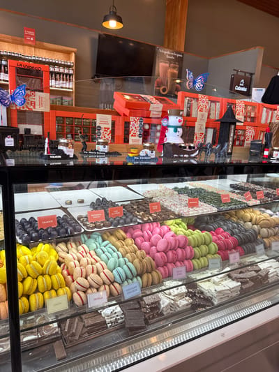 This detailed description pertains to the interior of a confectionery shop located in Tolhuin, Argentina.
The scene presents a well-lit retail environment, primarily focused on a multi-tiered glass display case filled with a variety of sweets. The case, positioned in the foreground, features three main levels of products. The top level contains rows of individually portioned chocolates and confections, identified by small orange labels reading "CALAFATE," "BANANITA DOCCA," and "CAFE ALCOHOL" among others. The middle two levels are densely packed with colorful macarons, arranged in large mounds by hue, including vibrant yellow ("LIMON"), light beige ("FRUTILLA"), light blue, pink ("FRUTOS ROJOS"), and light green ("PISTACHO"). The bottom shelf showcases an assortment of chocolate bars and fudge pieces, presented on white trays, varying in color from dark brown to white.
Behind the display case, a narrow dark counter serves as a decorative shelf. This shelf holds various miniature figures and items: a blue butterfly cutout, a miniature train engine labeled "TIERRA DEL FUEGO," small jars with location-specific labels such as "TOLHUIN," "EA. MARIA BEHETY," and "PTO. YAGHAN," a stylized black house labeled "USHUAIA," and a miniature snowman figure pulling a sleigh with reindeer, suggesting a festive or winter theme. Behind these decorations, multiple orange gift boxes, labeled "LA UNION CO." and bearing descriptive text like "BOMBONES SURTIDOS," are stacked vertically and horizontally.
The background features a rustic wood-paneled wall on the left, fitted with shelves holding numerous small glass bottles or jars. Above this, a red sign displays white text including "3 CUOTAS SIN INTERES." Centrally, a large wall-mounted flat-screen television displays an image of a beverage in a glass. To the right, a gray wall section features a hanging sign for "Cabrales" and a dark garment suspended from a hook. No individuals are visible within the frame. The overall environment suggests a well-maintained commercial space dedicated to the sale of specialty food items.