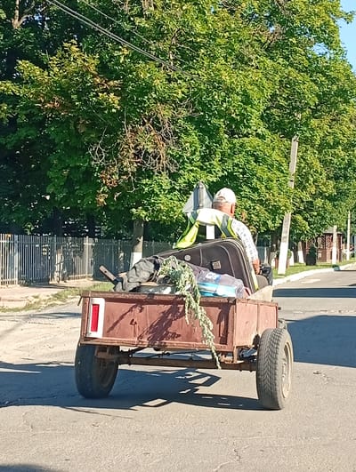The image shows a man wearing a high-visibility vest and a white cap driving a trailer with a load of items. The trailer is attached to a vehicle that is not fully visible. The items in the trailer include what appears to be luggage, plastic bottles, and some greenery. The man is facing away from the camera. The scene is outdoors on a paved road, with trees and a fence visible in the background. The overall impression is of a sunny day. The location is identified as Buțeni, Moldova.