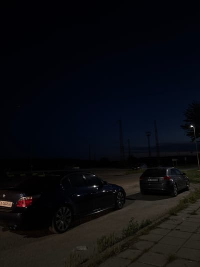 In the city of Dalgopol, Bulgaria, at night, two cars are parked on a dirt road next to a paved area. The car on the left is a dark-colored BMW sedan, seen from its rear quarter, parked at an angle. Its taillights are illuminated. The car on the right is a grey Audi station wagon, parked facing away from the viewer. Its license plate is visible and reads "B 4850 XB". Beyond the cars, industrial-looking towers with lights are visible against a dark sky. A single streetlamp illuminates a portion of the paved area in the foreground. The environment appears to be an outdoor, possibly semi-rural or industrial area, with sparse vegetation growing along the edge of the pavement. The sky is dark, indicating it is either late evening or early morning.
