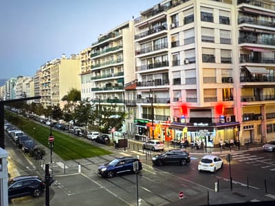 The image depicts a street scene in a European city, likely Nice, France, given the style of architecture and signage, not Porrentruy, Switzerland. In the foreground, a dark blue station wagon drives on tram tracks. To its right, a dark gray SUV is also on the street. Further up the street, a white hatchback turns at an intersection. Across the street, several people are seated at tables outside a cafe labeled "Le Muguet" and "BAR TABAC Presse". The buildings lining the street are multi-story, with balconies and awnings. On the left side of the image, a green median strip separates the tram tracks from a row of parked cars. The sky is overcast.