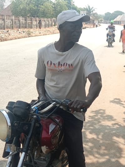In the city of Nassarawan Eggon, Nigeria, an adult male is observed positioned behind a motorcycle on a paved road under natural daylight. The male, appearing middle-aged or elderly with dark skin, wears a light-colored baseball cap, a cream-colored t-shirt bearing "Oxbow" in orange and red text, and dark trousers. His right hand grips the right handlebar of the motorcycle, and his head is turned to his right, looking off-frame. The motorcycle features a dark red/maroon fuel tank with visible paint wear and a chrome-bezeled round headlight. In the mid-ground, another individual on a motorcycle travels in the opposing direction, wearing a light shirt and a bright green head covering. To the right of this second motorcyclist, a pedestrian in an orange garment walks away from the viewer. The background features a low, weathered concrete wall paralleling the road, sparse dry vegetation, and distant trees. Two additional figures are visible walking near the wall in the left background. Shadows are cast on the road surface, consistent with direct sunlight.