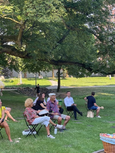 The image depicts an outdoor gathering in a park-like setting, likely in Stupava, Slovakia, under the shade of a large, mature tree. Several individuals are seated on chairs, facing to the right. In the foreground, on the left, a person wearing a yellow dress is visible from the waist down, with bare feet on the grass. Next to them, a woman with blonde hair and glasses is seated, wearing a patterned top and white pants. To her right, an elderly man with a white cap and a plaid shirt is seated, looking towards the right. Behind them, other individuals are seated, including a woman in a black dress and a man in a white shirt. Further back, a person in a dark blue shirt and denim shorts is seated. The environment consists of a well-maintained grassy area with stone borders and some scattered trees. In the background, there are stone walls and structures. A picnic basket with fruit is partially visible in the bottom right corner. The lighting suggests it is daytime, possibly late afternoon, with dappled sunlight filtering through the tree canopy.
