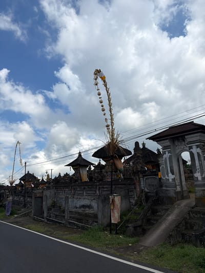 In the city of Susut, Indonesia, a view of a temple complex on the side of a road is captured under a partly cloudy sky. The temple features dark, multi-tiered roofs and elaborate stone walls, with a large, ornate archway visible on the right. Two tall, decorative bamboo poles, known as "Penjor," stand prominently in front of the temple. One Penjor, adorned with offerings and streamers, arches high into the sky on the right side of the frame, while another shorter one is visible on the left. The foreground shows a paved road with a white line marking its edge, and a strip of green grass separates the road from the temple grounds. The overall scene depicts a traditional Balinese temple entrance against a backdrop of a dynamic sky.
