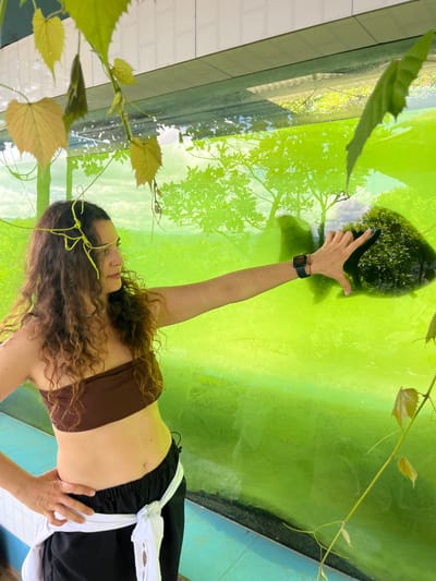 An adult female with curly brown hair is standing in front of a large, bright green glass enclosure. She is wearing a brown strapless top and black pants with a white sash tied around her waist. Her left arm is bent at the elbow, with her hand on her hip. Her right arm is extended towards the glass, with her hand touching the surface. The glass reflects a scene of trees and sky. The enclosure appears to be some kind of display or habitat. The floor is tiled in blue and white. Green vines with leaves hang into the frame from the top of the image. The scene is likely an outdoor or semi-outdoor location.