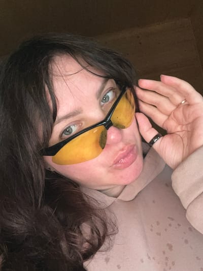 This close-up portrait, captured in Protasy, Russia, features an adult female posing for the camera. Her face is angled upwards and tilted, with her gaze directed forward. She has dark hair, light-colored eyes (likely blue or green), and full, glossy lips that are slightly parted.
She is wearing distinctive glasses with dark frames and unconventional yellow-orange, semi-translucent lenses that appear to have a textured or irregular surface. Her left hand is raised towards her head, with a thin, gold-toned ring on her ring finger and a thicker, dark-colored ring on her middle finger. She is dressed in a light-colored, possibly beige or pale pink, hooded sweatshirt or sweater, which shows several scattered dark spots or stains on its fabric.
The background is a dark, featureless surface, suggesting an indoor or confined setting. The lighting illuminates her face, highlighting her features. Her expression is composed and direct, indicating a posed shot. There is no visible text in the image.