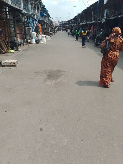 A street scene depicts a narrow road lined with shops and people. The road is grey and stretches into the distance where more people are visible. On either side, buildings, partially constructed from wood and metal, provide shelter for shops. A few people are walking along the street. A woman in a patterned dress stands to the right, her back to the viewer. A motorcyclist is further down the road. The street appears busy.